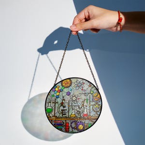 May include: A round, stained-glass window with a black metal chain. The window features a colorful laboratory scene with beakers, test tubes, and scientific equipment. The window is held up by a hand against a white background.