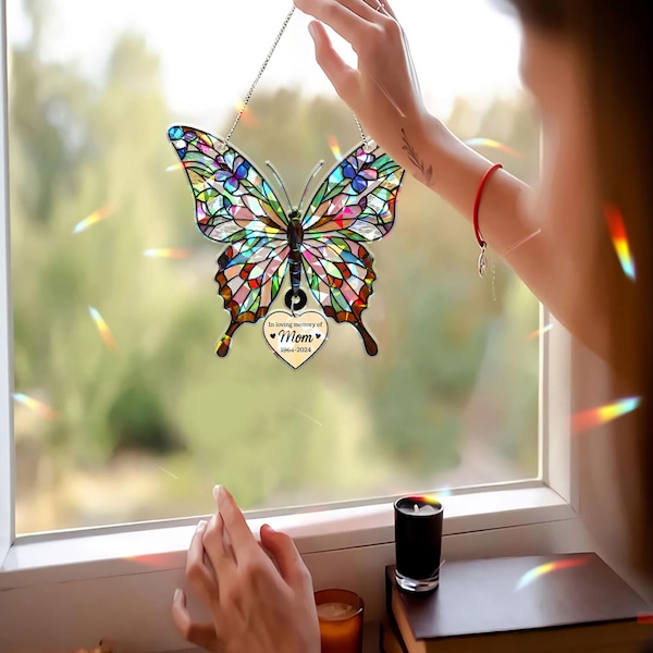 May include: A stained glass butterfly suncatcher with multicolored segments and a hanging heart-shaped charm. The charm features the text "In loving memory of Mom 1964-2024" with two small black hearts. The ornament is suspended by a thin metal chain.