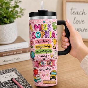 May include: A pink tumbler with a black lid, handle, and straw, featuring the text MISS SOPHIA and school-themed graphics including a pencil, globe, trophy, apple, books, and a yellow school bus, with the phrase teaching inspiring encouraging making a DIFFERENCE every day.