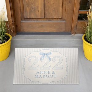 May include: A rectangular doormat with a light blue and white striped background. The mat features a light blue decorative frame with the numbers "222" and the names "ANNE & MARGOT" in a matching light blue font. A light blue bow is at the top.