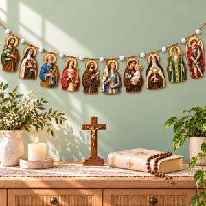 May include: A decorative garland featuring ten individual wooden cutouts of religious figures, each with a gold halo, strung together with white beads on a brown cord. The figures are depicted in colorful robes and traditional attire.