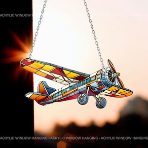 Puede incluir: Un avión de estilo vitral para colgar en la ventana. El avión es rojo, amarillo, azul y blanco. Está colgado de una cadena plateada.