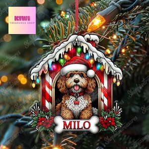 Pode incluir: Um enfeite de Natal de madeira com um cão usando um chapéu de Papai Noel dentro de uma casinha de cachorro. A casinha é decorada com listras vermelhas e brancas, luzes e o nome "MILO" em uma etiqueta em forma de osso. O enfeite está pendurado em uma árvore de Natal.