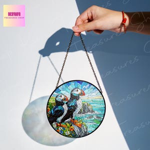 Puede incluir: Una ventana redonda de vidrio tintado colgante presenta un dise&ntilde;o de mosaico de tres frailecillos en un acantilado con olas oce&aacute;nicas. Los frailecillos son azules, blancos y naranjas. El fondo es un mosaico de azul, verde y blanco.