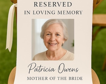 May include: A cream-colored sign with the text "RESERVED IN LOVING MEMORY" and a photo of a smiling woman. Below the photo, it reads "Patricia Owens MOTHER OF THE BRIDE". The sign is attached to a wooden chair with cream-colored ribbons.