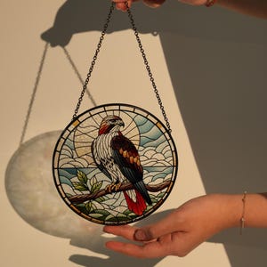 May include: A stained glass window hanging ornament featuring a red-tailed hawk perched on a branch. The hawk is depicted in a realistic style with detailed feathers and a bright red tail. The background is a blue sky with white clouds.