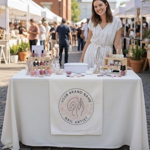 Puede incluir: Un camino de mesa blanco con un logotipo circular que incluye el texto YOUR BRAND NAME NAIL ARTIST y una ilustraci&oacute;n lineal de una mano. La mesa est&aacute; dispuesta con varios frascos de esmalte de u&ntilde;as, pinceles y peque&ntilde;os recipientes.