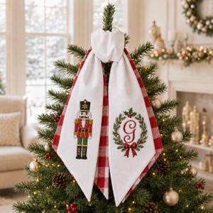 Peut inclure: Un cimier de sapin de Noël en tissu blanc avec deux longs pans pointus au motif à carreaux rouges et blancs sur l'envers. Un pan présente un casse-noisette brodé, tandis que l'autre arbore une couronne verte avec une lettre G rouge.