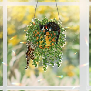 May include: A hanging stained glass cat suncatcher. The design features a black and white cat resting in a hanging planter with green leaves, and a tabby cat climbing up the side. The suncatcher is suspended by a silver chain.