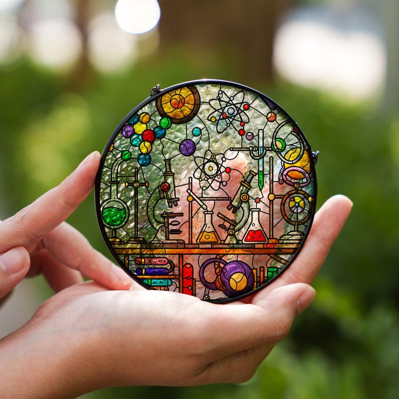 May include: A round stained glass panel featuring a science lab design. The artwork includes colorful beakers, microscopes, and atom models. The panel is held in front of a blurred green background, showcasing the intricate details and vibrant colors of the glass.