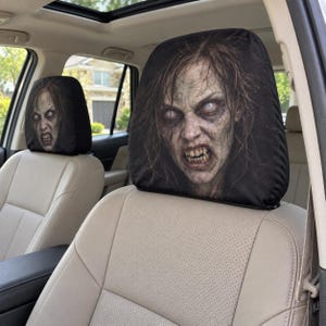 May include: A pair of black car headrest covers featuring a high-resolution, realistic graphic print of a pale, snarling zombie with dishevelled hair, sunken eyes, and bared teeth. The covers are fitted over the headrests of light beige car seats.