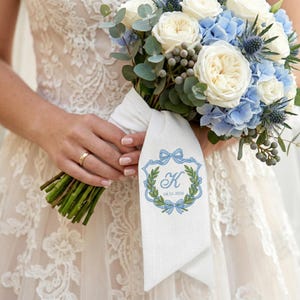 Op de afbeelding: Een bruidsboeket met witte rozen, blauwe hortensia's en groene eucalyptus, omwikkeld met een wit stoffen lint. Het lint toont een geborduurd blauw embleem met de letter K en de datum 04.10.2026 in blauwe draad.