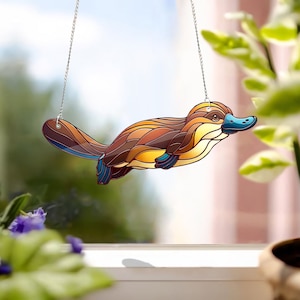 May include: A stained glass hanging ornament of a platypus with brown, yellow, and blue colors. The platypus is hanging from a silver chain.