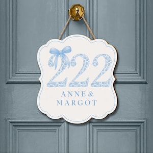 May include: A white scalloped wall sign with a light blue border, hanging from a jute rope. The sign features the number 222 in a light blue floral pattern with a light blue bow on the first digit, and the text ANNE & MARGOT in light blue.
