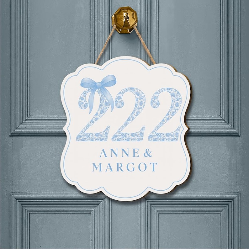 May include: A white scalloped wall sign with a light blue border, hanging from a jute rope. The sign features the number 222 in a light blue floral pattern with a light blue bow on the first digit, and the text ANNE & MARGOT in light blue.