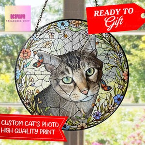 Puede incluir: Un atrapasoles circular de vidriera con el retrato detallado de un gato atigrado de ojos verdes. El gato está rodeado por un borde decorativo de flores coloridas y dos mariposas. La pieza cuelga de una cadena de metal de color negro.