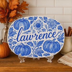 Könnte beinhalten: Eine ovale, weiße Keramikplatte mit blauem Blumen- und Kürbismuster. Das Wort "Lawrence" ist kursiv in der Mitte geschrieben. Die Platte wird auf einem transparenten Ständer präsentiert, mit einem Kürbis und Herbstlaub im Hintergrund.