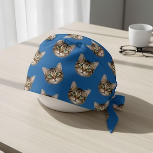 May include: A blue surgical scrub cap featuring a repeating pattern of cat faces with green eyes and brown and tan fur. The cap has a tie in the back and is sitting on a white bowl.