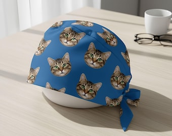 May include: A blue surgical scrub cap with a repeating pattern of cat faces. The cats have green eyes and brown and tan fur. The cap has a tie at the back and is on a white bowl.