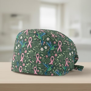 May include: A green scrub cap featuring a pattern of pink ribbons, hummingbirds, and white flowers. The cap is designed with a tie in the back for an adjustable fit. The fabric has a soft, matte finish.