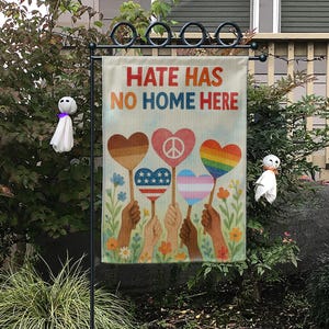 May include: A garden flag with the text "HATE HAS NO HOME HERE" in orange and red. The flag features hands holding heart-shaped designs with a peace sign, rainbow, and American flag. The flag is displayed outdoors.