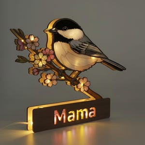 Puede incluir: Lámpara decorativa de madera con forma de pájaro en una rama florecida. La base de la lámpara tiene la palabra "Mama" iluminada. El pájaro es negro, blanco y gris, con flores rosas y blancas. Con cable de alimentación.