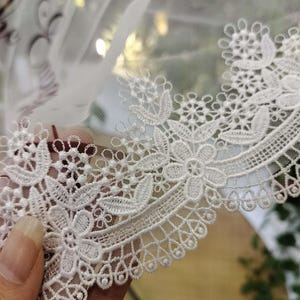 May include: A white tulle veil featuring an intricate floral lace border with scalloped edges. The sheer fabric displays dark text partially visible at the top, while the embroidery showcases detailed flower and leaf patterns in white thread.