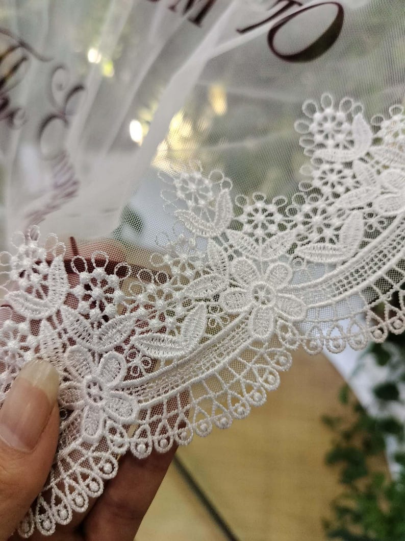 May include: A white tulle veil featuring an intricate floral lace border with scalloped edges. The sheer fabric displays dark text partially visible at the top, while the embroidery showcases detailed flower and leaf patterns in white thread.