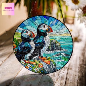 Puede incluir: Una ventana de vitral colgada de una cadena, que representa dos frailecillos posados en un acantilado con vistas a un oc&eacute;ano azul con olas. Los frailecillos est&aacute;n hechos de piezas de vidrio de colores, con colores azul, negro, blanco y naranja. El fondo presenta un cielo azul con nubes y una costa rocosa.