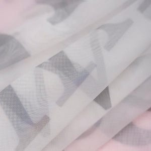 May include: A sheer, semi-transparent white fabric featuring a repeating pattern of dark gray, bold, block-style capital letters. The material is folded, creating soft, layered pleats that obscure parts of the printed text throughout the surface.