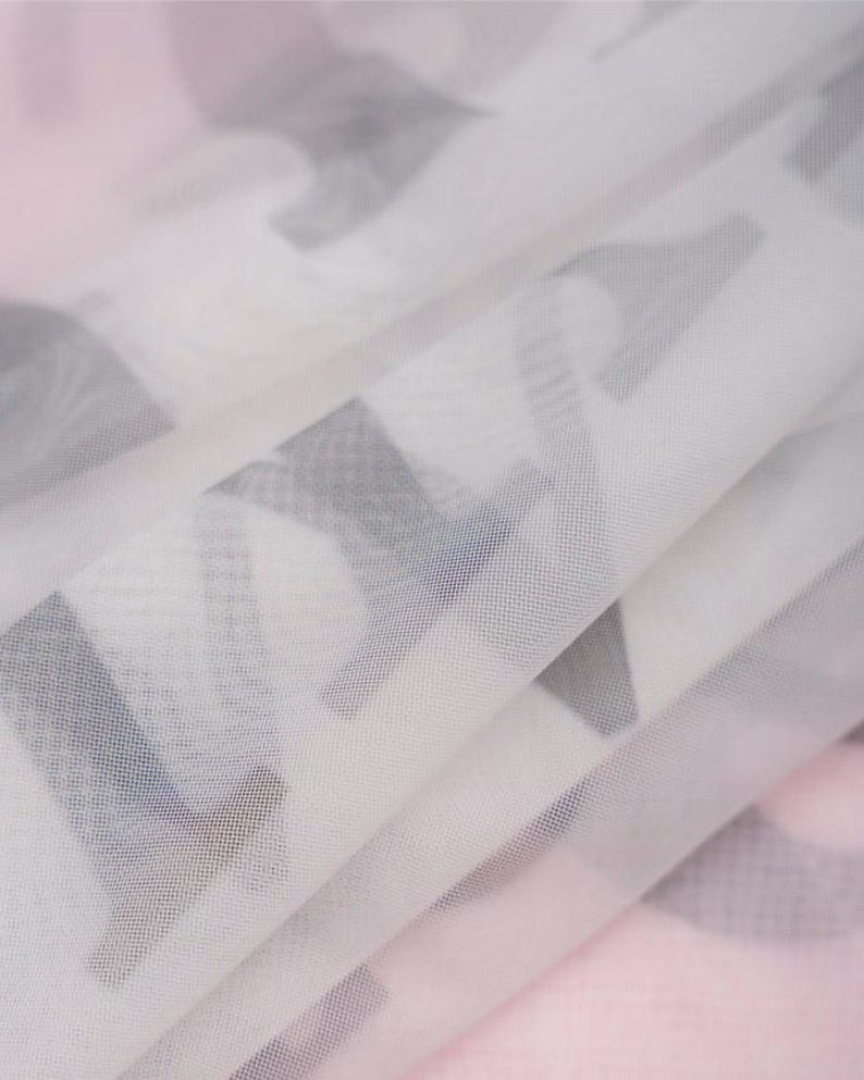 May include: A sheer, semi-transparent white fabric featuring a repeating pattern of dark gray, bold, block-style capital letters. The material is folded, creating soft, layered pleats that obscure parts of the printed text throughout the surface.