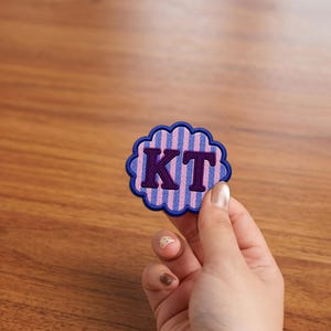 May include: A scalloped circular embroidered patch featuring vertical light pink and blue stripes with the dark purple capital letters KT centered on the front. The patch has a thick dark blue stitched border around the entire outer edge.