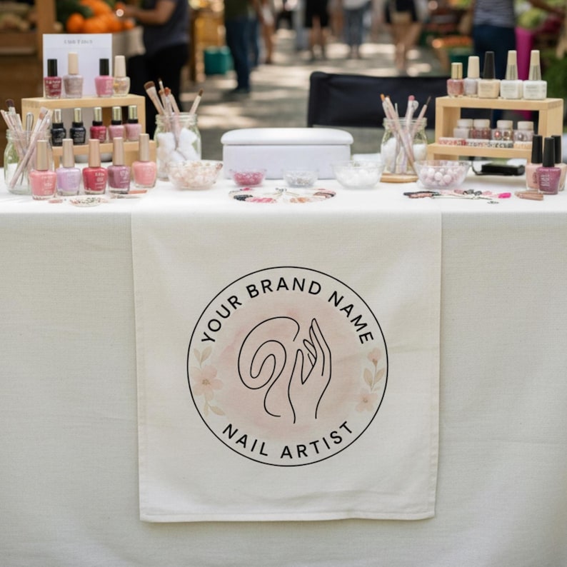 Puede incluir: Un camino de mesa de tela blanca con un logotipo circular que incluye el texto YOUR BRAND NAME NAIL ARTIST y una ilustraci&oacute;n lineal de una mano. El expositor incluye soportes de madera escalonados con varios esmaltes y pinceles.