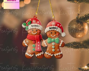 May include: Two gingerbread man Christmas ornaments. Each ornament is wearing a Santa hat and has a name on the hat. One has a red scarf and the other has a green bow tie. The ornaments are hanging from gold strings.