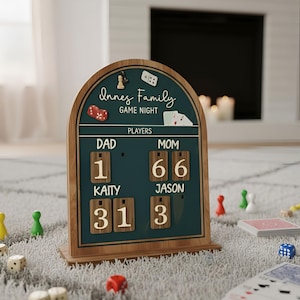 May include: Wooden game night scoreboard with the text "Innes Family Game Night." The scoreboard has a teal background with wooden numbers and player names. Dice, playing cards, and game pieces are scattered around the board.