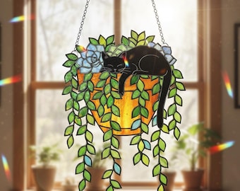 May include: Stained glass hanging planter with a black cat sleeping among green leaves and blue flowers. The planter is a warm orange color and is suspended by a chain. The leaves have green and blue accents.