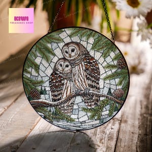May include: A round stained glass window hanging with a chain. The window features a mosaic design of two owls perched on a branch with pine needles and snow. The owls are brown and white with large, round eyes.