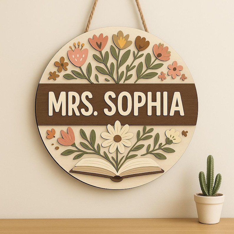 May include: A round wooden sign with the name "MRS. SOPHIA" in white letters on a brown band. The sign is decorated with floral and leaf cutouts in shades of pink, yellow, and green. A book is at the bottom. A rope is attached for hanging.