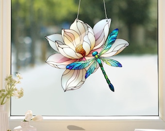 May include: A stained glass window hanging features a white magnolia flower with a dragonfly perched on it. The dragonfly has blue, green, and yellow wings.
