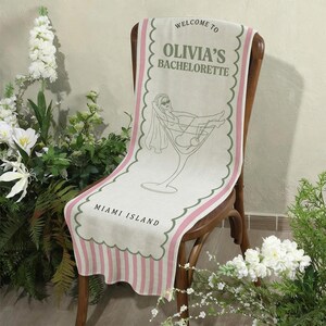 May include: A linen table runner draped over a wooden chair. The runner features the text "WELCOME TO OLIVIA'S BACHELORETTE MIAMI ISLAND" with a cocktail glass illustration. The design includes pink and green accents.