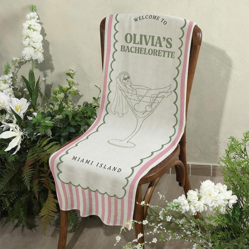 May include: A linen table runner draped over a wooden chair. The runner features the text "WELCOME TO OLIVIA'S BACHELORETTE MIAMI ISLAND" with a cocktail glass illustration. The design includes pink and green accents.