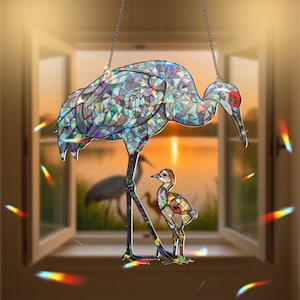 May include: A stained glass hanging ornament featuring a pair of iridescent cranes. The larger crane is standing with its legs outstretched, while the smaller crane is standing next to it. The background is a blurry image of a sunset over a lake.
