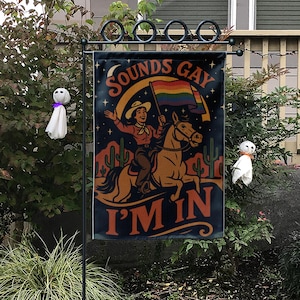 May include: A garden flag with the text "SOUNDS GAY I'M IN" features a cowgirl riding a horse and holding a rainbow flag. The flag has a dark blue background with a desert scene and stars. Two small ghost decorations are on either side.