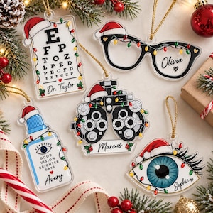 May include: Five clear acrylic Christmas ornaments featuring eye-themed designs: an eye chart with Dr. Taylor, black glasses with Olivia, a phoropter with Marcus, an eye drop bottle with Avery, and a blue eye with Sophie. Each has a Santa hat and lights.