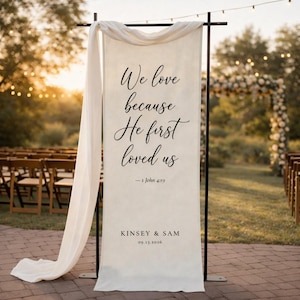 Puede incluir: Una pancarta de boda color crema con letras negras que dice "We love because He first loved us" y "- 1 John 4:19". La pancarta tambi&eacute;n incluye los nombres "KINSEY & SAM" y la fecha "09.13.2026". La pancarta est&aacute; drapeada con una tela blanca.
