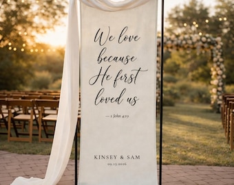 May include: A cream-colored wedding banner with black script that reads "We love because He first loved us" and "- 1 John 4:19". The banner also includes the names "KINSEY & SAM" and the date "09.13.2026". The banner is draped with a white fabric.