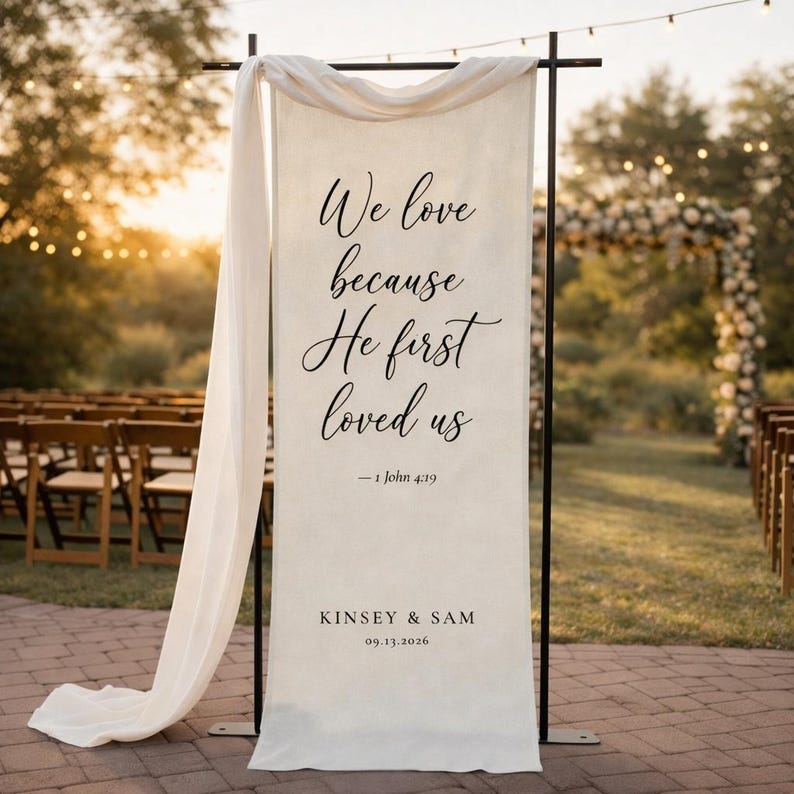 Puede incluir: Una pancarta de boda color crema con letras negras que dice "We love because He first loved us" y "- 1 John 4:19". La pancarta tambi&eacute;n incluye los nombres "KINSEY & SAM" y la fecha "09.13.2026". La pancarta est&aacute; drapeada con una tela blanca.