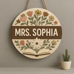 May include: A round wooden sign with the name "MRS. SOPHIA" in white lettering. The sign features a floral design with flowers in shades of peach, yellow, and brown, and a book illustration. A jute rope is attached for hanging.