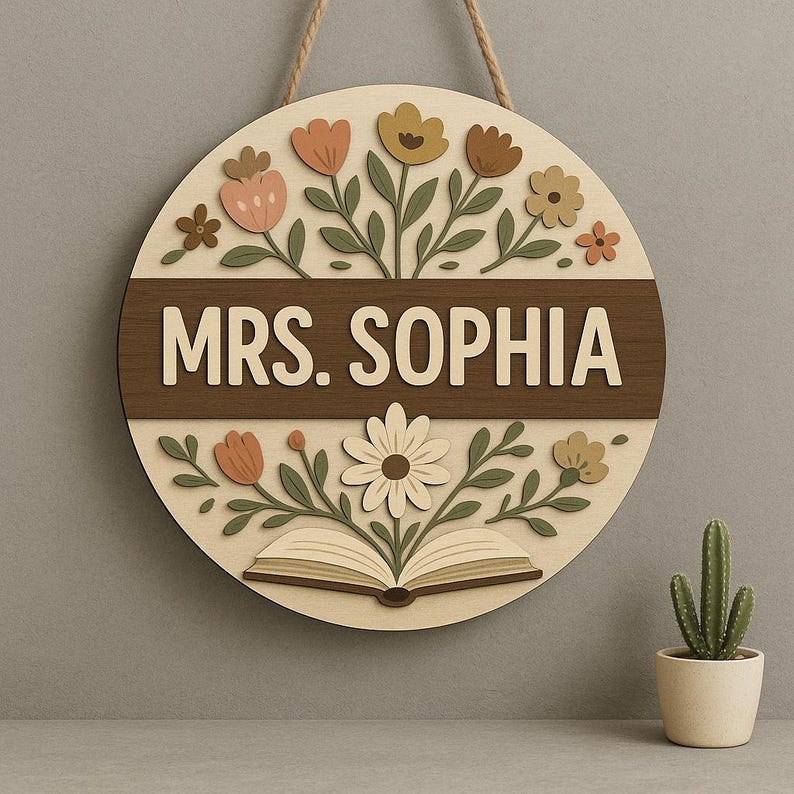 May include: A round wooden sign with the name "MRS. SOPHIA" in white lettering. The sign features a floral design with flowers in shades of peach, yellow, and brown, and a book illustration. A jute rope is attached for hanging.