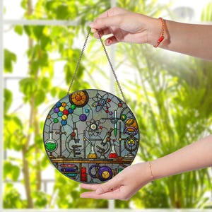 May include: A round, stained glass window hanging with a science theme. The design features beakers, microscopes, atoms, and colorful spheres. The window is held by a chain and is displayed in front of a window with green foliage.